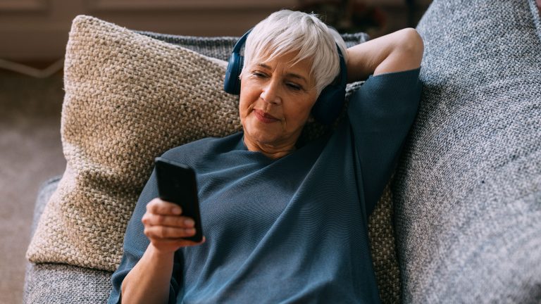 Eine Person liegt auf dem Sofa und hört über Kopfhörer etwas an ihrem Handy auf das sie schaut.