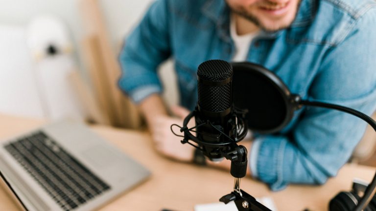 Der Fokus liegt auf einem Podcastmikrofon im Vordergrund hinter der eine Person mit Laptop an einem Tisch sitzt.