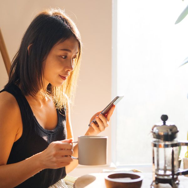 Eine Person sitzt mit einem iPhone in der einen Hand an einem Tisch, auf dem eine French Press steht; in der anderen Hand hält sie eine Kaffeetasse.