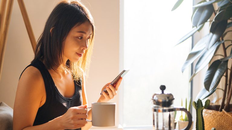 Eine Person sitzt mit einem iPhone in der einen Hand an einem Tisch, auf dem eine French Press steht; in der anderen Hand hält sie eine Kaffeetasse.