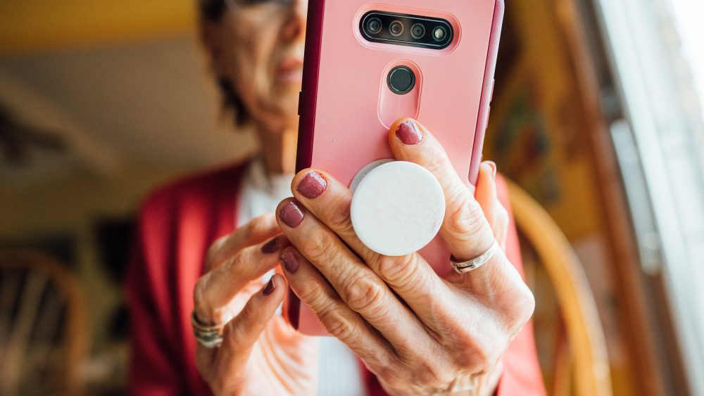 Eine Person hält ein Smartphone mithilfe eines PopSockets in der Hand. Der Fokus der Aufnahme liegt auf der Rückseite des Smartphones sowie den Händen der Person.