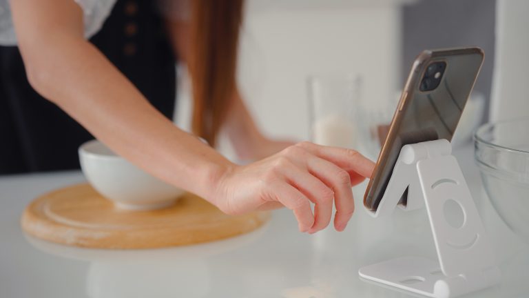 Ein Smartphone steht auf einem Standfuß auf einer Arbeitsfläche in einer Küche. Im Hintergrund ist eine weiße Schüssel auf einem Holzbrett zu sehen.