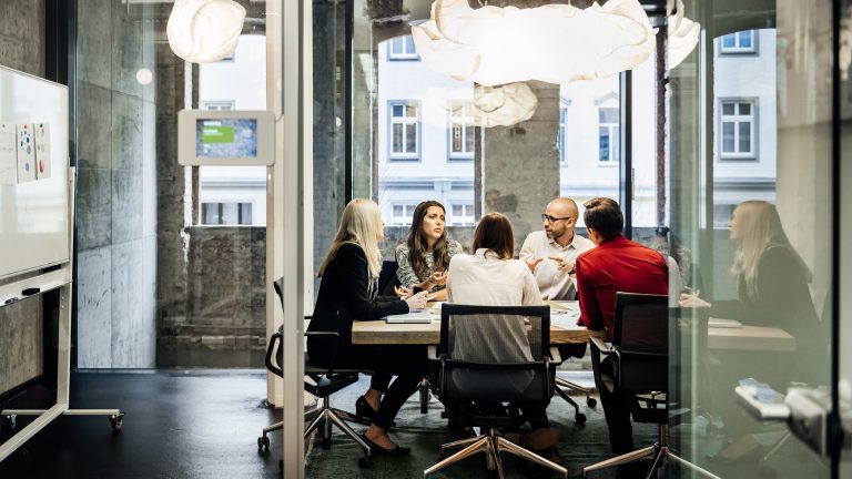 Eine Gruppe von Personen sitzt in einem Meeting-Raum in einem industriell wirkenden Bürogebäude.