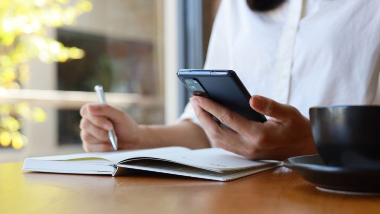 Eine Person sitzt mit einem Stift vor einem Notizblock und hält ihr Smartphone in der anderen Hand.