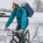 Eine Person mit einem Rucksack auf dem Rücken ist bei Schnee mit einem Fahrrad unterwegs.