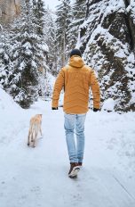 Eine Person läuft auf einem schneebedeckten Waldweg mit einem Hund spazieren und ist dabei winterlich gekleidet.