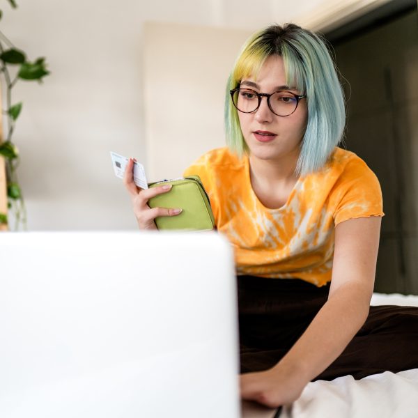 Eine Person sitzt auf einem Bett vor dem geöffneten Laptop. In der Hand hält sie ein Portemonnaie und eine Kreditkarte.