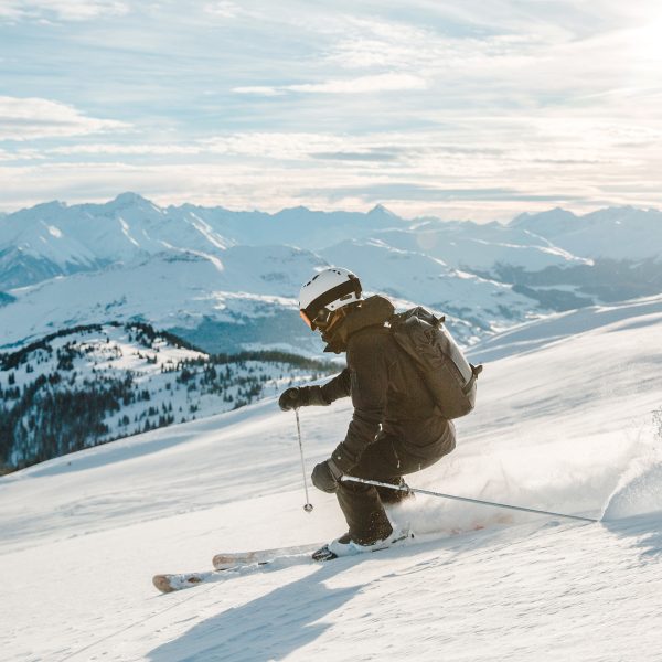 Eine Person fährt auf Skiern die Piste herunter. Im Hintergrund sind schneebedeckte Berge zu sehen.